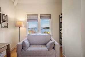 Une pièce avec un canapé gris, une lampe, une bibliothèque et une fenêtre donnant sur la mer.