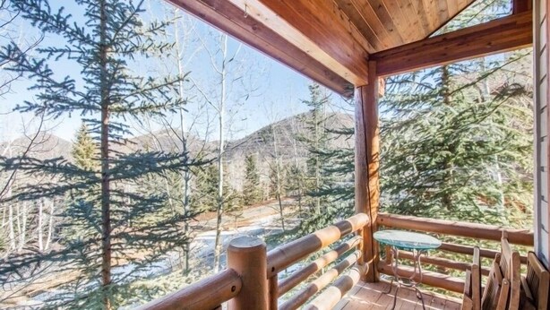 A wooden deck with a glass table and chairs, overlooking a forested area with tall trees and mountains in the background.