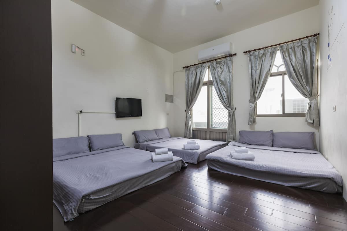 A room with three single beds, a flat-screen TV mounted on the wall, and a window with curtains.