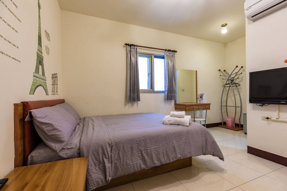 A bedroom with a bed, a wooden nightstand, a window with curtains, a television, and a wall-mounted mirror.