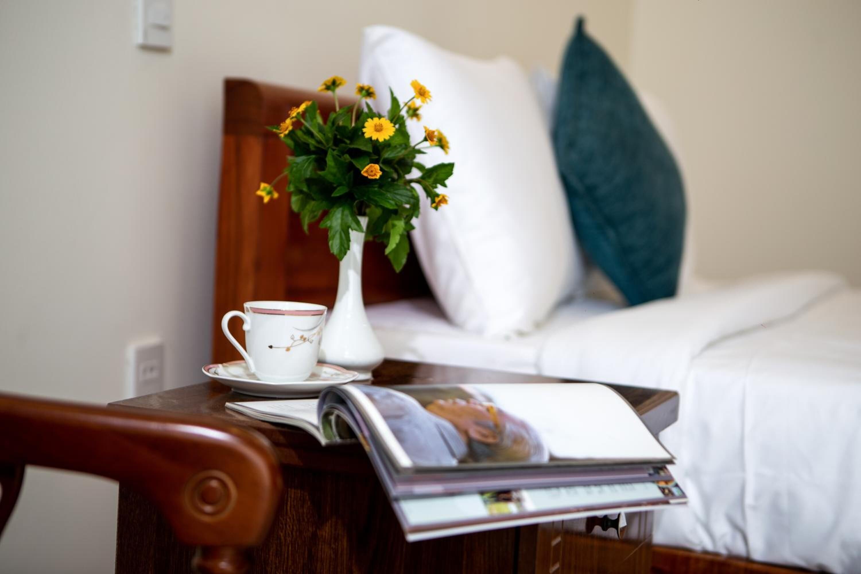 A hotel room with a wooden nightstand, a white vase with yellow flowers, a white cup, and a magazine.