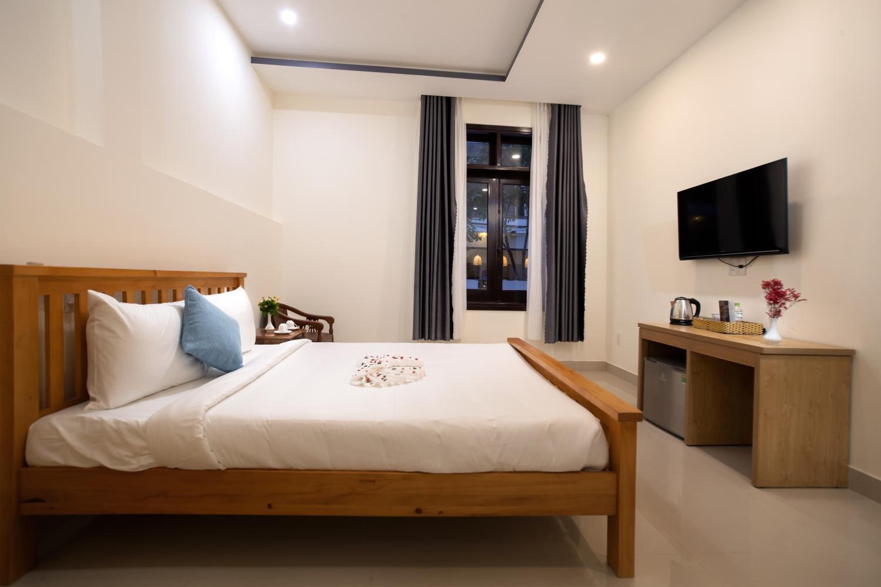 A neatly arranged bedroom with a wooden bed, a small bedside table, a television, and a window with curtains.