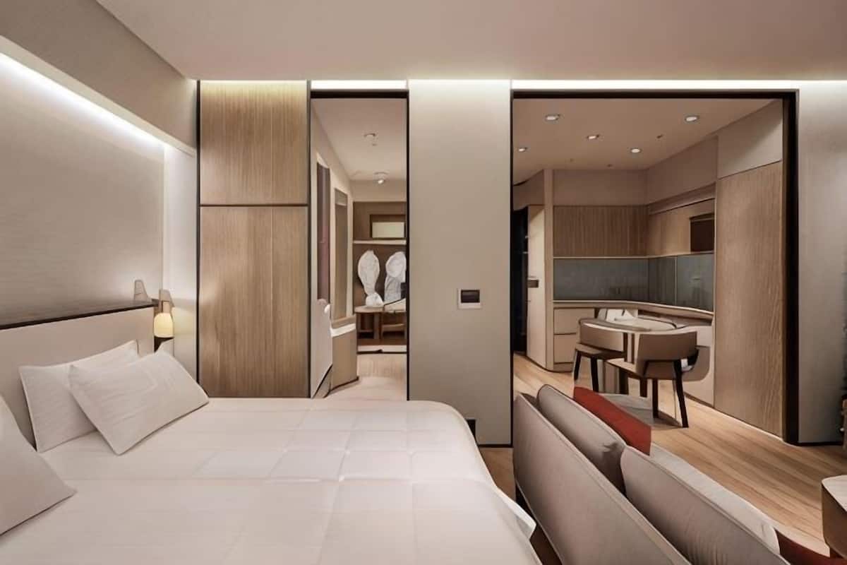 Une chambre d’hôtel moderne dotée d’un grand lit, d’un coin salon séparé, d’une kitchenette et d’une salle de bain visible à travers une porte ouverte.