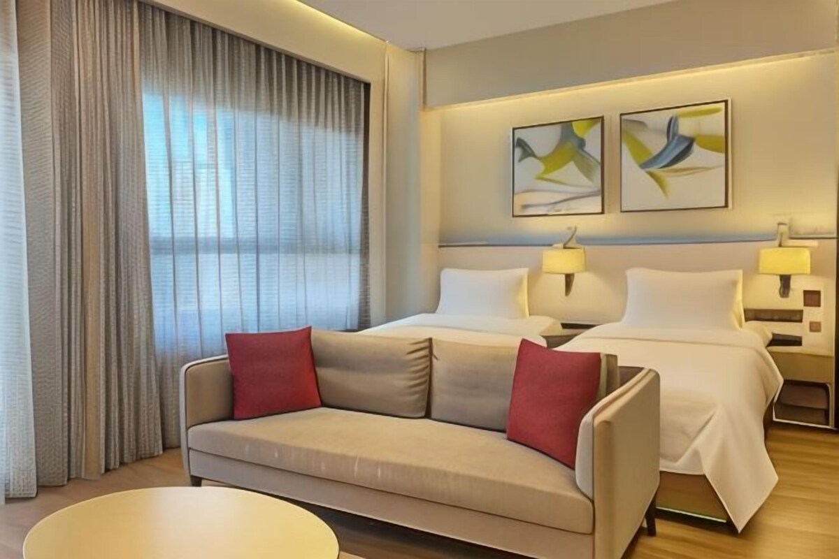 Habitación de hotel con una cama, dos mesitas de noche con lámparas, un sofá con cojines rojos, una mesa de centro redonda y un ventanal con cortinas.
