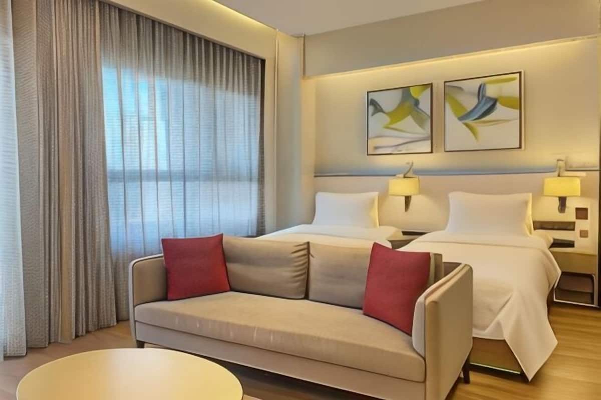 Une chambre d’hôtel avec un lit, deux tables de chevet avec des lampes, un canapé avec des coussins rouges, une table basse ronde et une grande fenêtre avec des rideaux.