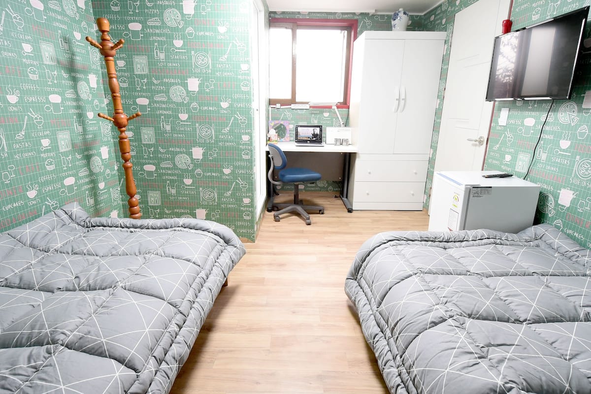 A bedroom with two beds, a desk with a chair, a television, and a wardrobe.