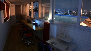 A room with a long table, several chairs, a clock on the wall, and large windows overlooking a cityscape at night. - Photo Park Guesthouse (Seoul)