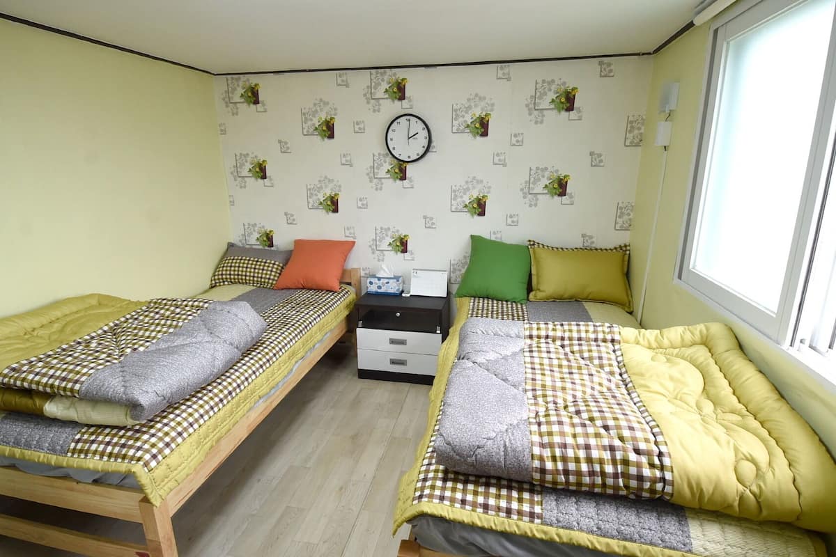 A bedroom with two single beds, a bedside table with a lamp, a clock on the wall, and a window providing natural light.