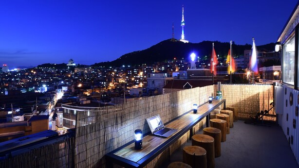 A rooftop bar with a laptop and lanterns, overlooking a cityscape with illuminated buildings and a tower. - Photo Park Guesthouse (Seoul)