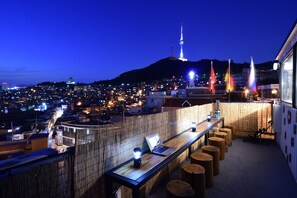 Un bar en la azotea con un ordenador portátil y faroles, con vistas a un paisaje urbano de edificios iluminados y una torre.