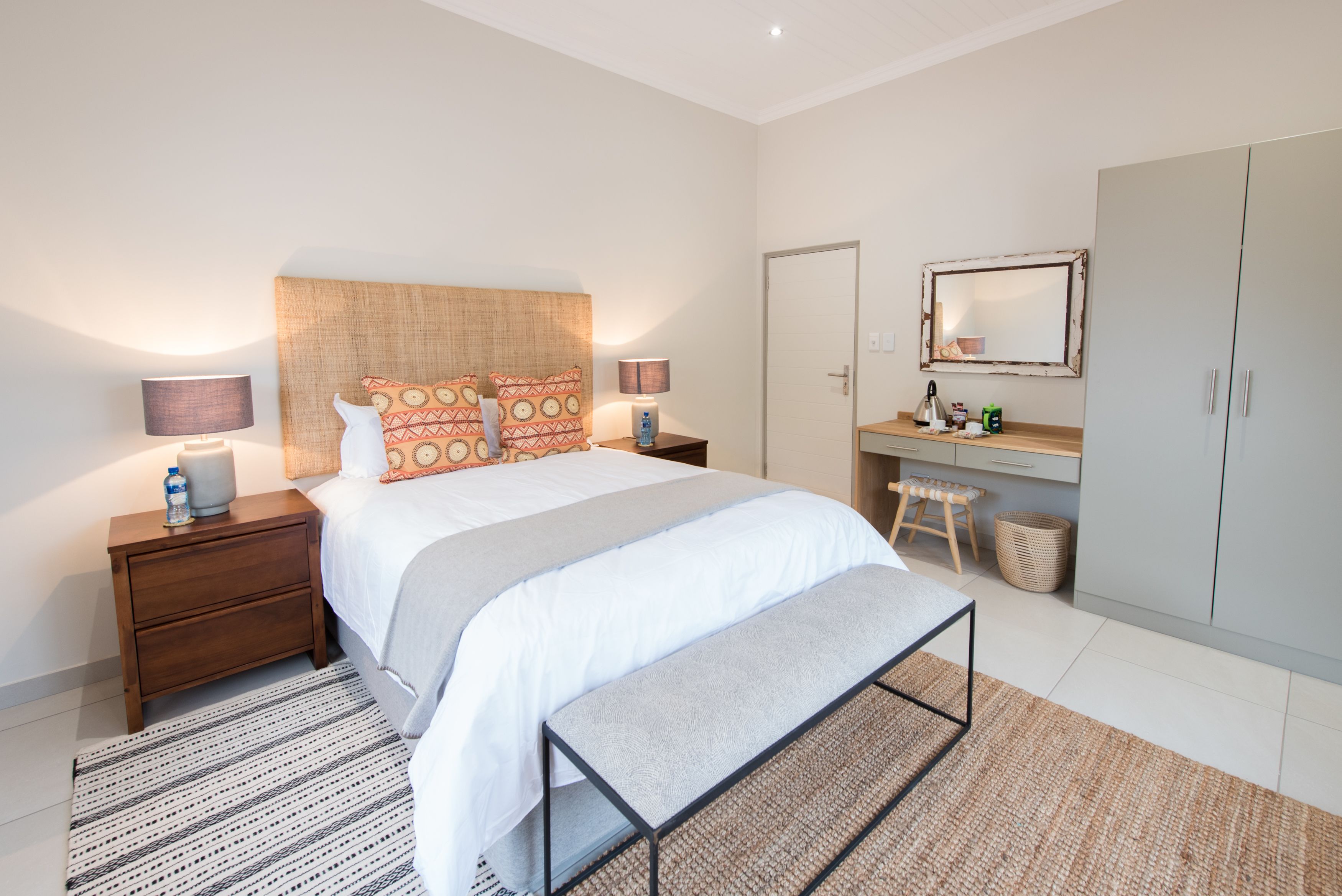 Un dormitorio moderno con una cama grande, mesitas de noche con lámparas, un banco, un armario y un espejo.