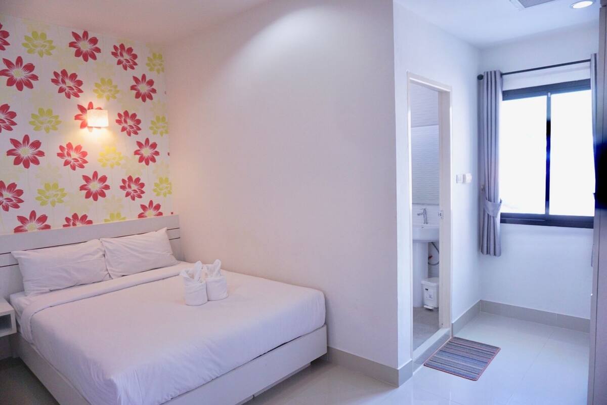 Habitación de hotel con cama, mesita de noche, lámpara de pared, baño visible a través de una puerta abierta y ventana con cortinas.