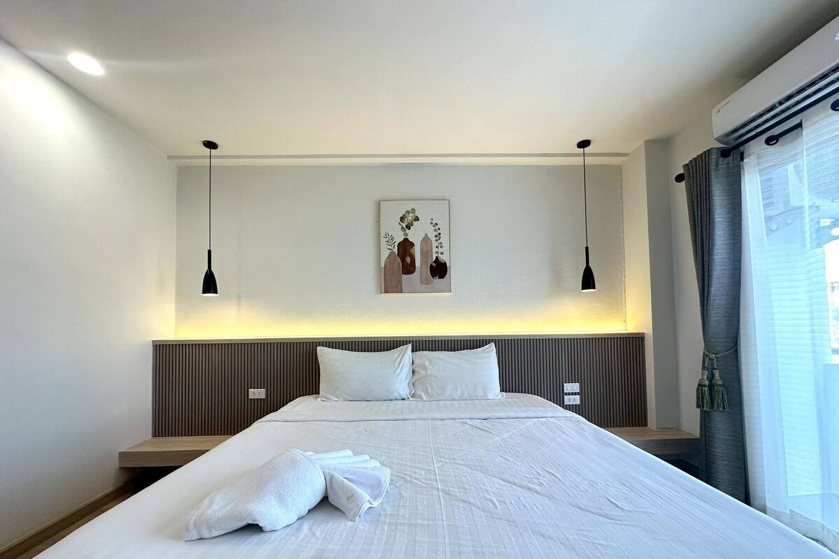 Una cama bien hecha con ropa de cama y toallas blancas, una obra de arte en la pared y un equipo de aire acondicionado instalado en el techo.