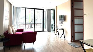 A modern living room with a red sofa, a small table, and a TV on a stand. There is a large window with curtains and a view of a construction site. - Bhundaree Richy (Hat Yai)