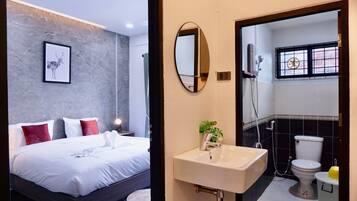 A modern hotel room with a large bed, a bedside table with a lamp, a framed picture on the wall, and a bathroom with a sink, mirror, toilet, and shower.