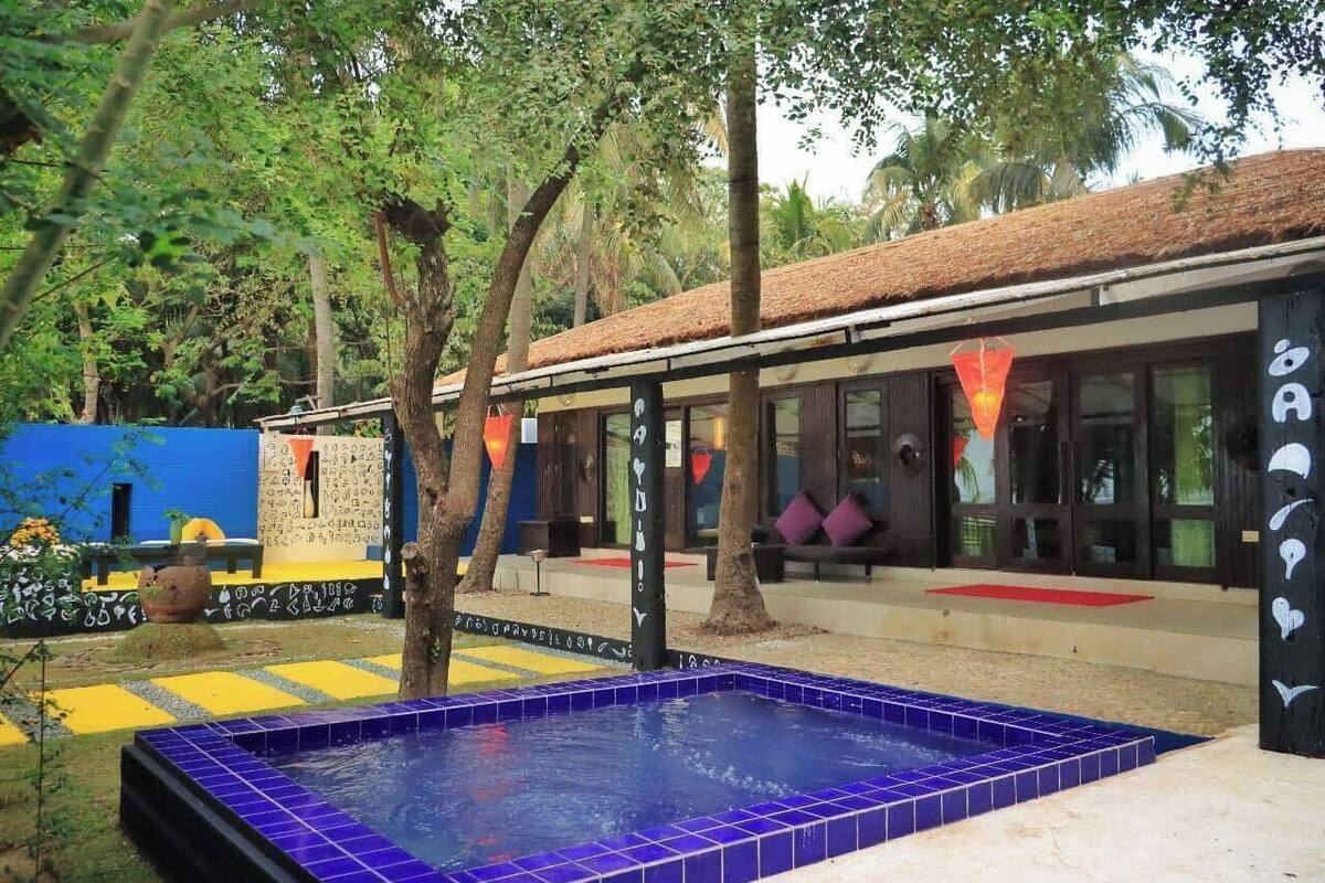 A pool area with a blue tiled border, surrounded by trees and a building with large windows and a thatched roof.