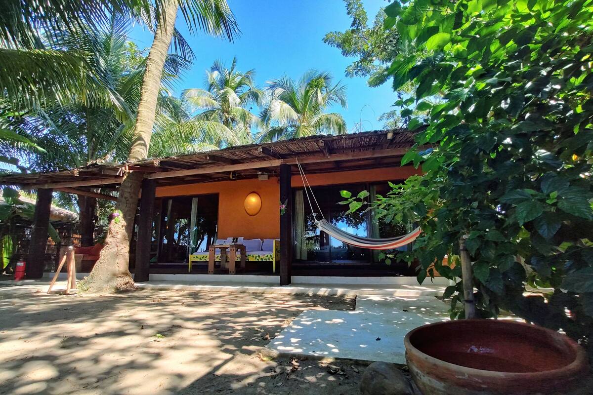 A rustic building with a thatched roof, a hammock, and a large window overlooking a garden with palm trees.