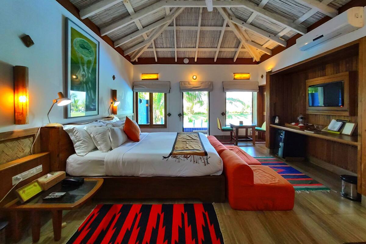 A spacious room with a large bed, a desk, and a television. There are windows providing natural light and a view of the outdoors.