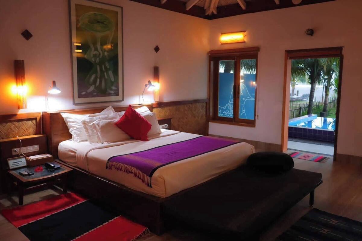 A hotel room with a bed, bedside tables, a nightstand, a window with a view of palm trees, and a painting on the wall.