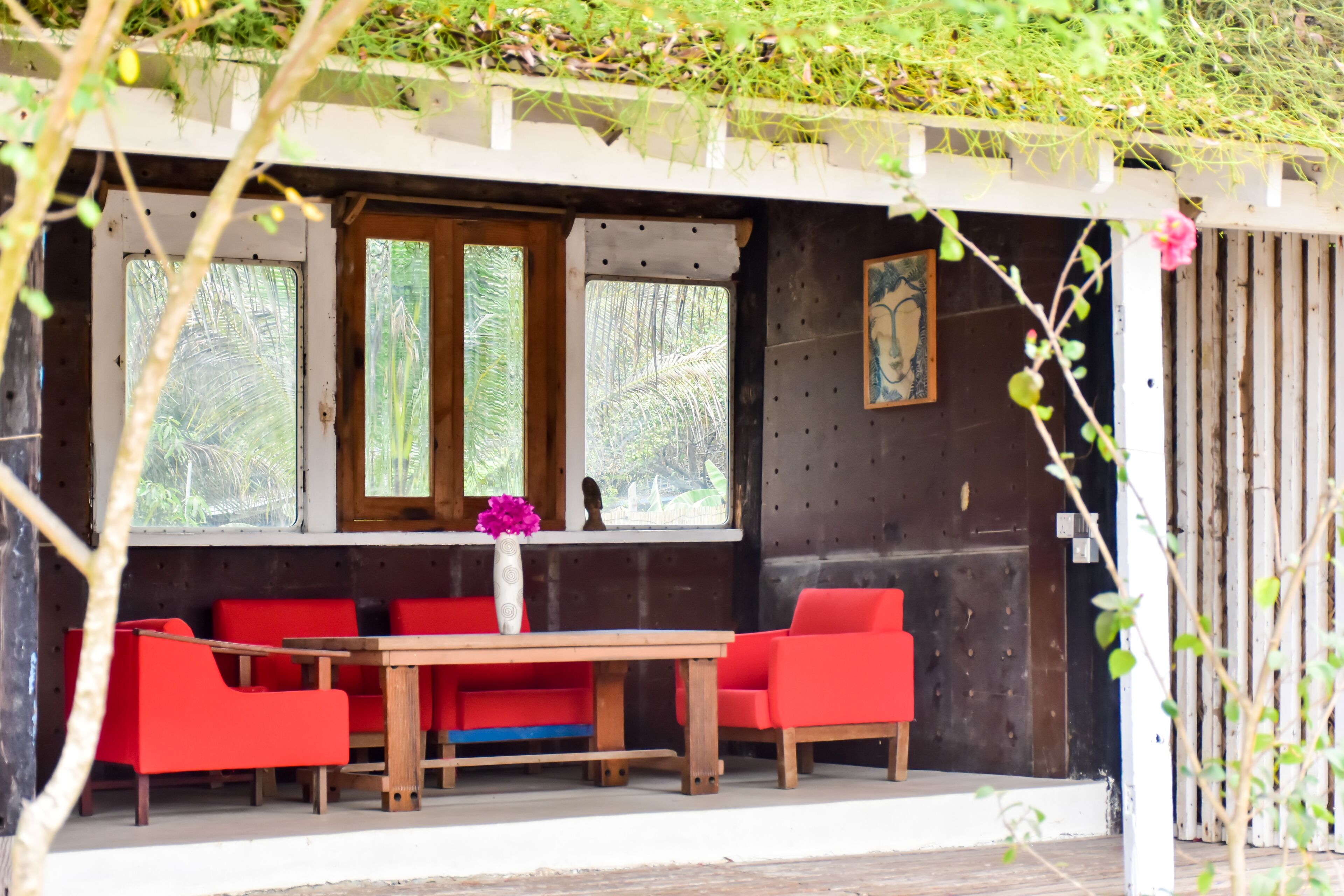 A room with a wooden table and red chairs, large windows with a view of greenery, and a painting on the wall.