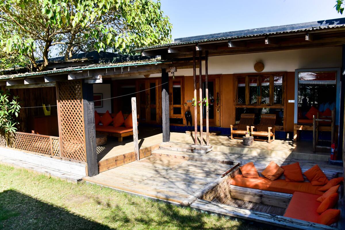 A covered outdoor seating area with wooden furniture and orange cushions, a wooden fence, and a tree providing shade.