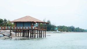 A wooden structure with a tiled roof, elevated on stilts over water, featuring a balcony with glass railings. - Mermaid Beach Resort (Cox's Bazar)