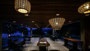 An outdoor dining area with a long table set for a meal, surrounded by chairs, under hanging wicker lamps, with a view of the sea and trees in the background. - Cala Laiya (San Juan)