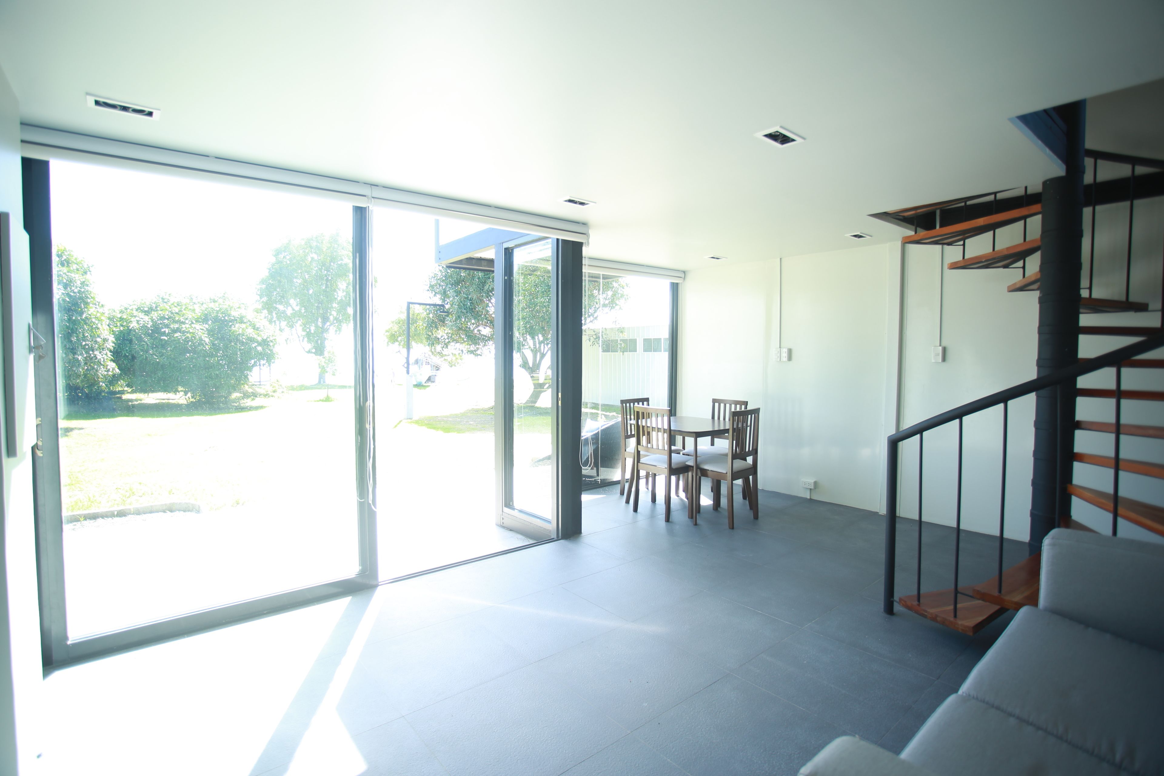 A spacious room with a large sliding glass door leading to an outdoor area, a staircase with a wooden finish, and a dining area with chairs and a table.