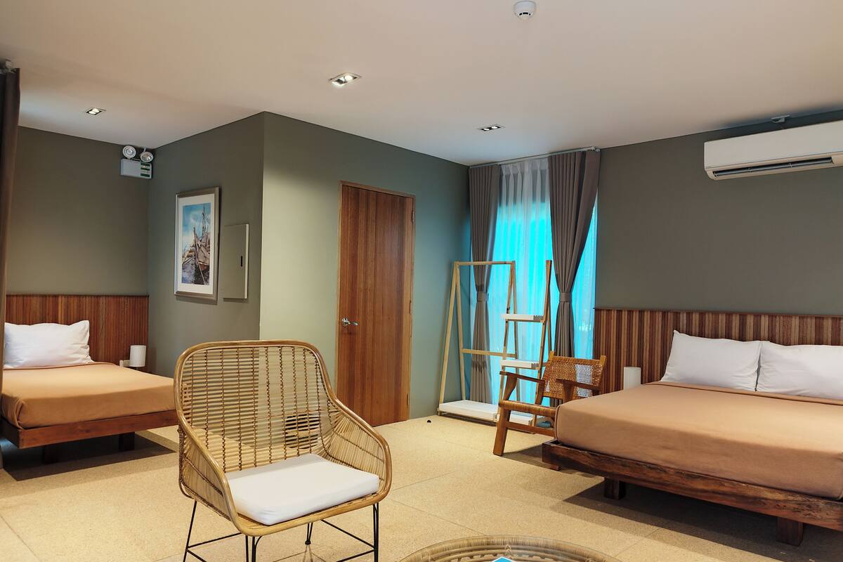 A hotel room with two single beds, a wicker chair, a wooden ladder, and a ceiling-mounted air conditioner.