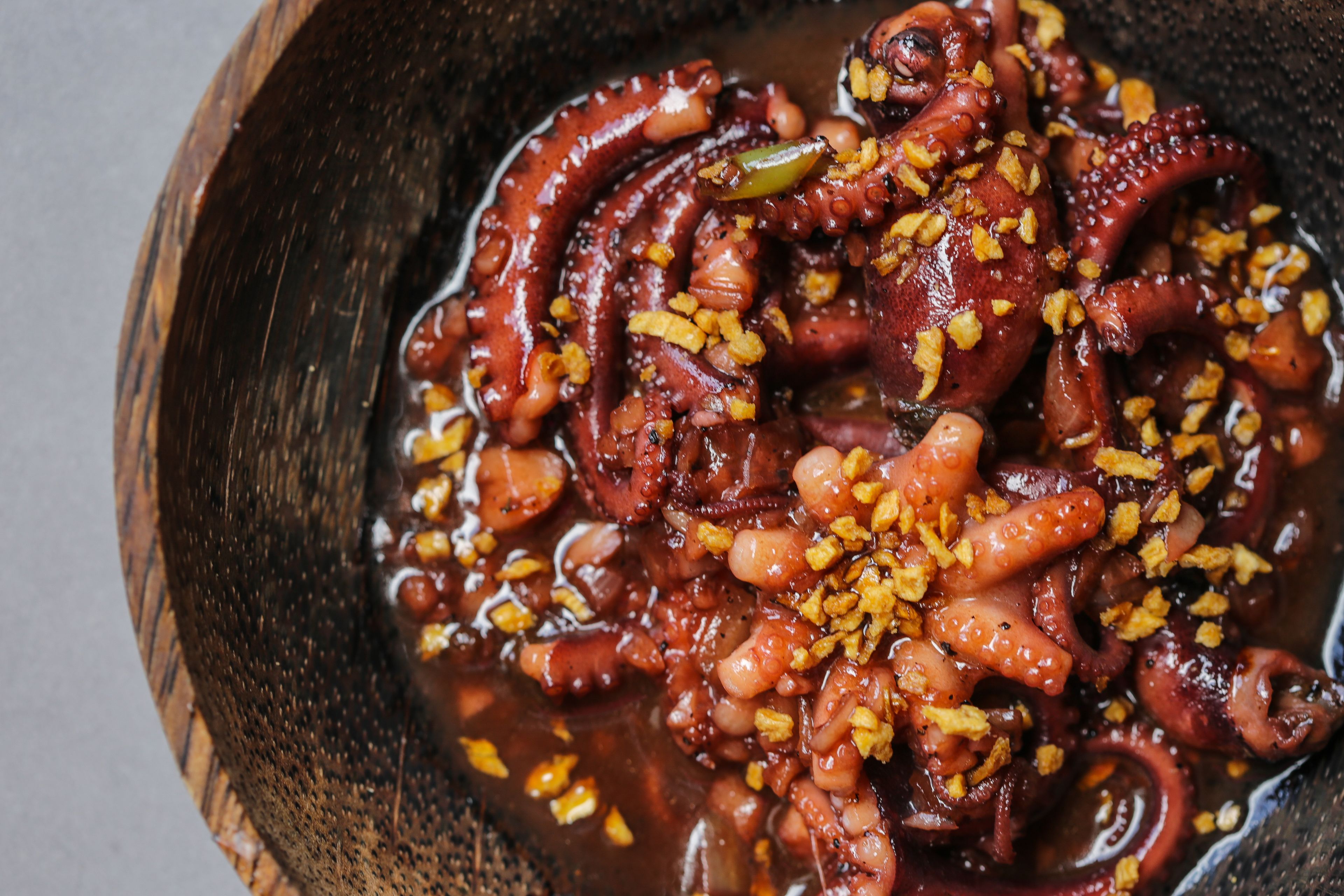 A wooden bowl containing octopus with a sauce and garnished with crispy bits.