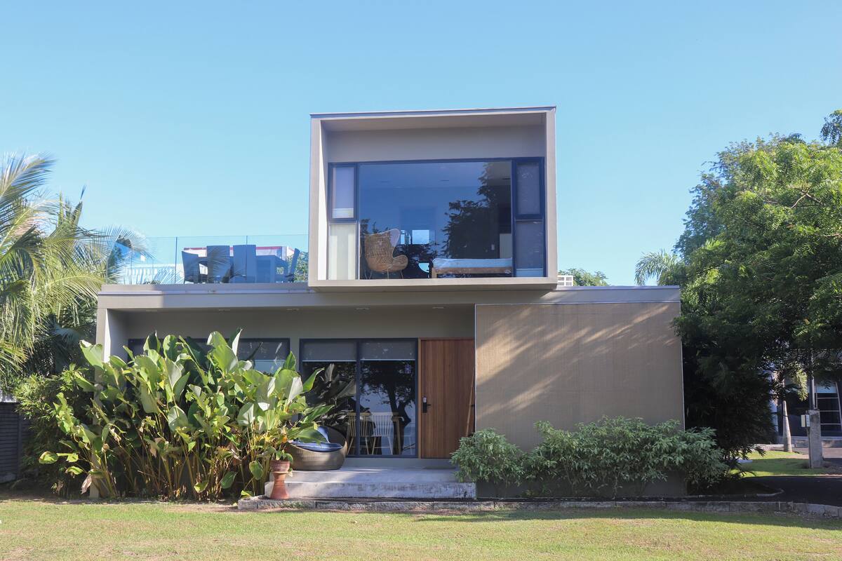 A modern two-story house with a large glass window on the upper floor, a wooden door, and a small porch area with a chair and table.