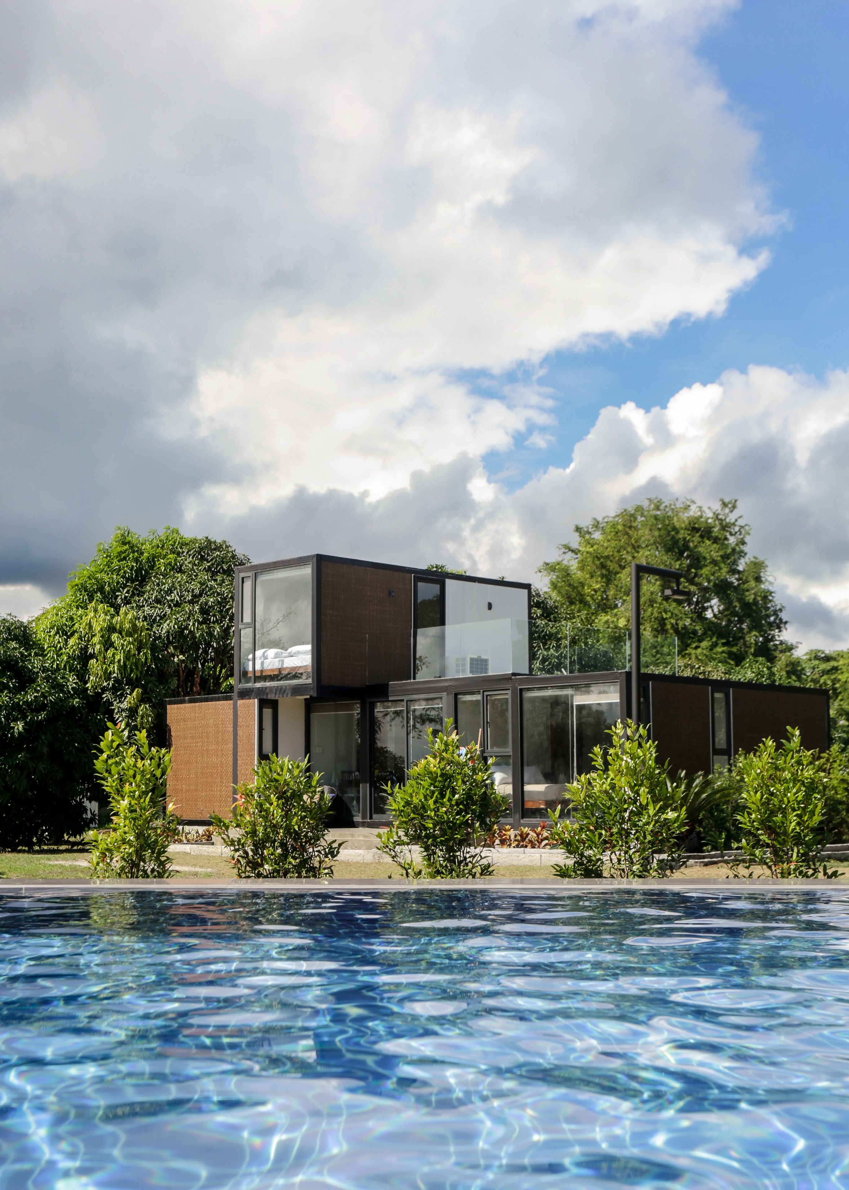 A modern building with a glass facade, a swimming pool in the foreground, and lush greenery in the background.