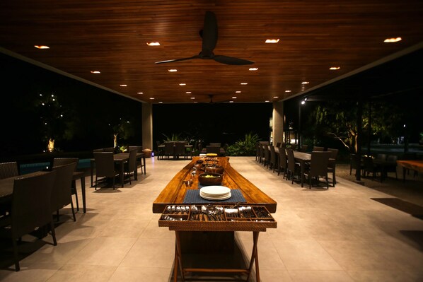 A spacious outdoor dining area with a long wooden table set for multiple people, surrounded by chairs, and a ceiling fan overhead. - Cala Laiya (San Juan)