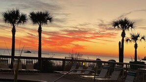 A poolside area with lounge chairs and palm trees, overlooking the ocean at sunset.