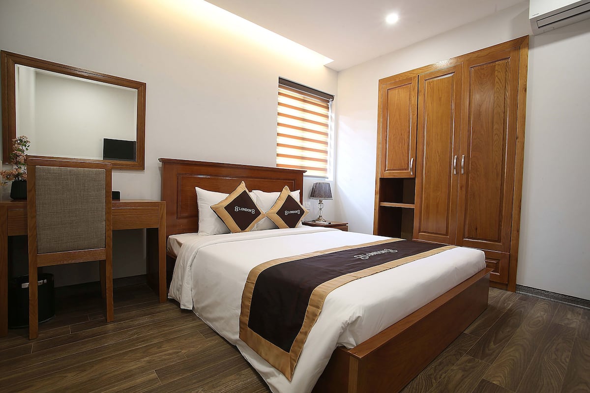 A bedroom with a bed, wooden wardrobe, a desk with a chair, a mirror, and a window with blinds.