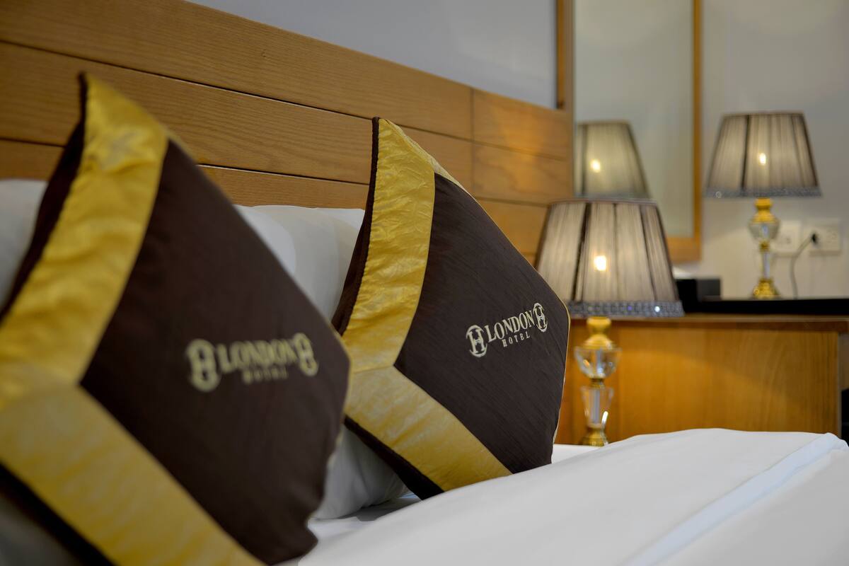 A hotel bed with pillows featuring the hotel's logo, a bedside lamp, and a wooden headboard.