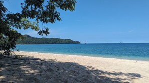 A beach with clear water, a sandy shore, and a forested island in the distance.