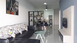 Una sala de estar moderna con un sofá negro, una mesa de centro de vidrio y un televisor montado en la pared. Al fondo, se observa un comedor con una mesa y sillas.