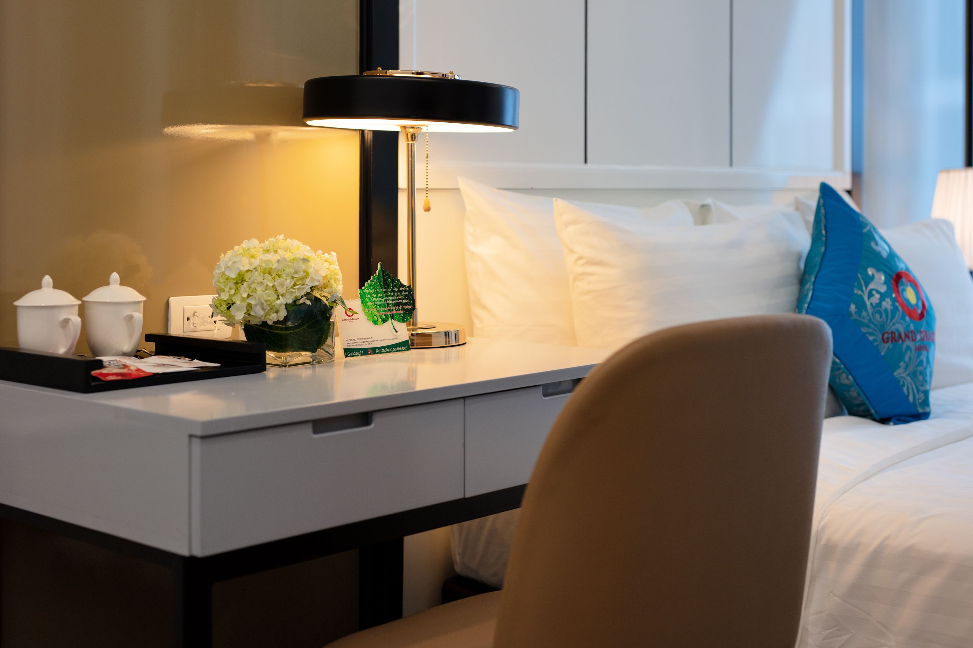Ein Hotelzimmer mit einem ordentlich bezogenen Bett, einem Schreibtisch mit Lampe, einer Blumenvase und einem blauen Kissen.