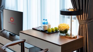 Een hotelkamer met een bureau, een televisie waarop het logo van het hotel te zien is, een dienblad met appels, waterflesjes en een vaas met bloemen.