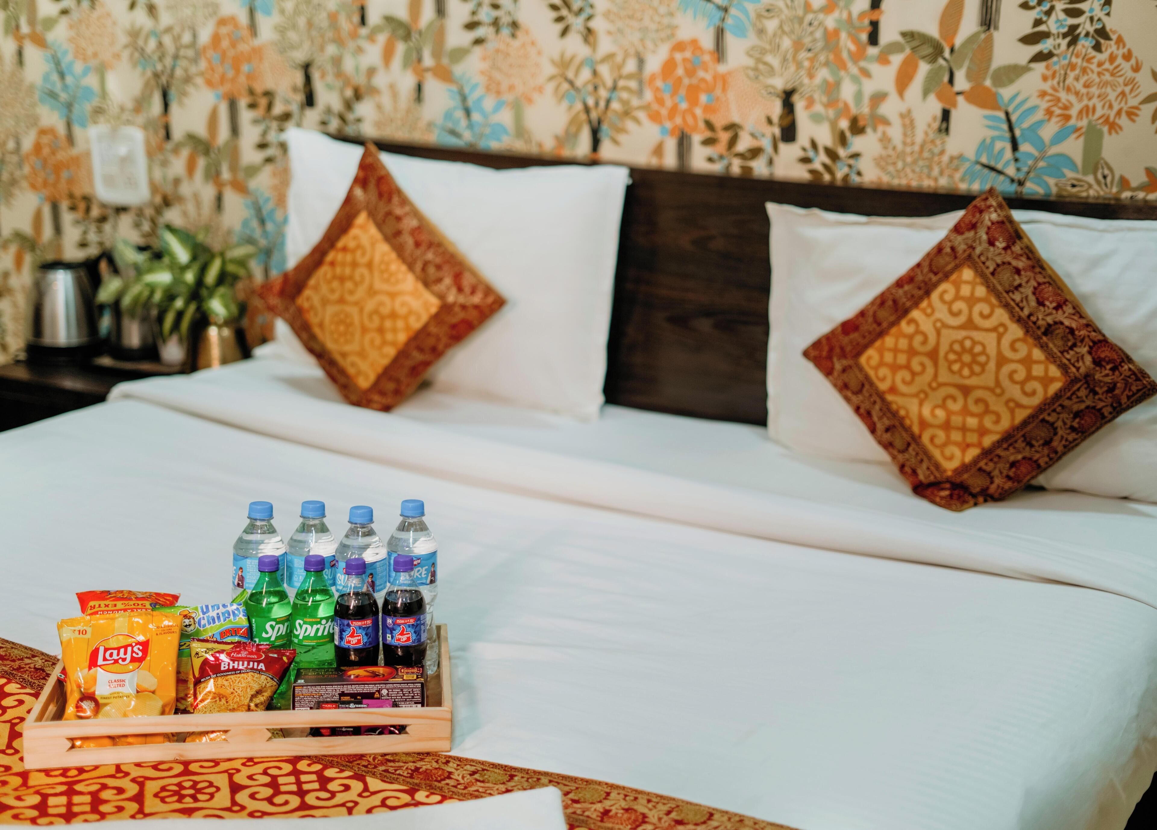 Una cama bien hecha con sábanas blancas y almohadas decorativas, una bandeja con aperitivos y bebidas, y un fondo de papel tapiz con motivos florales.