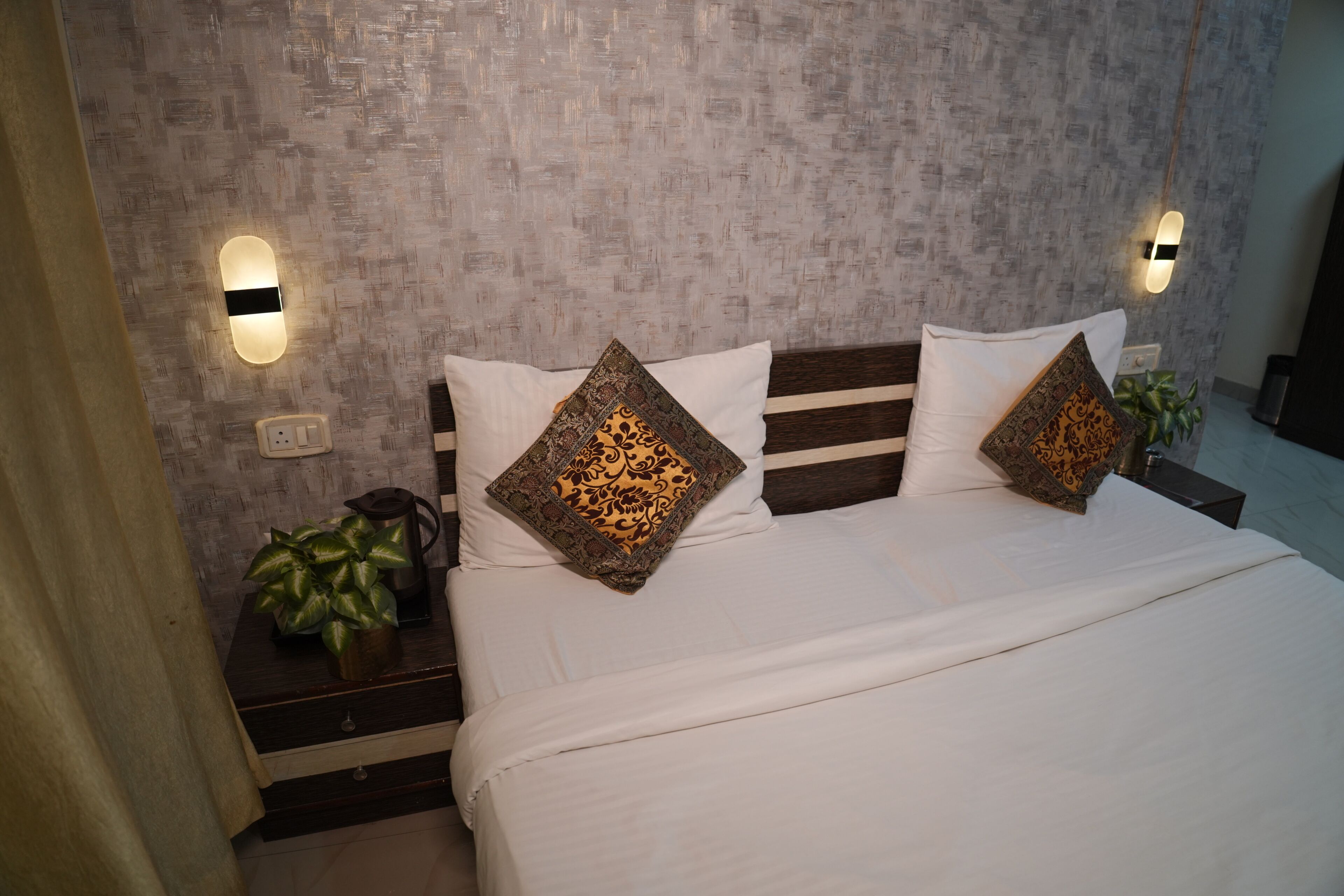 A neatly made bed with a striped headboard, two decorative pillows, a bedside plant, and wall-mounted light fixtures.
