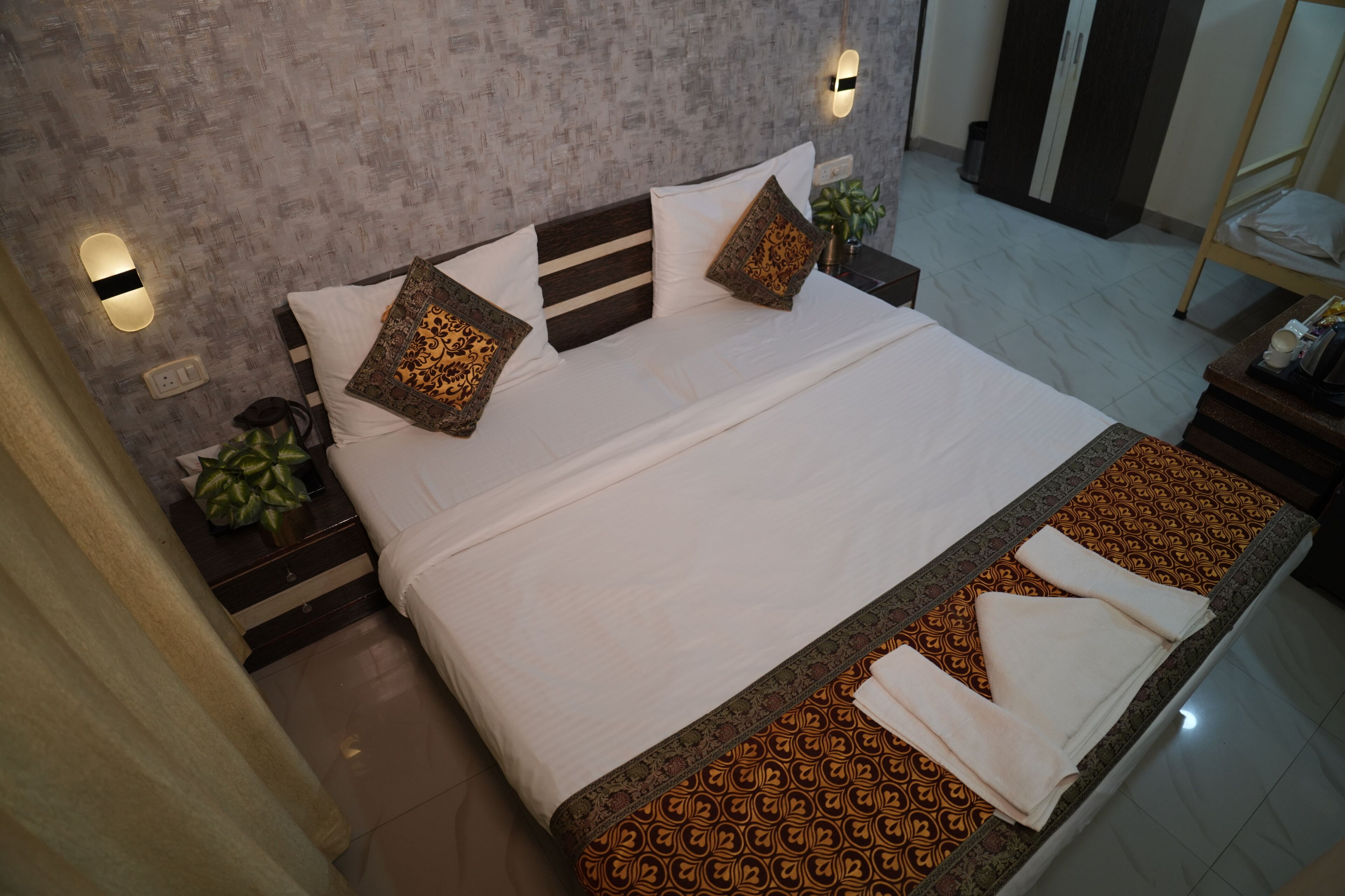 Una cama bien hecha con colcha y almohadas estampadas, una mesita de noche con una planta, y una lámpara de pared.
