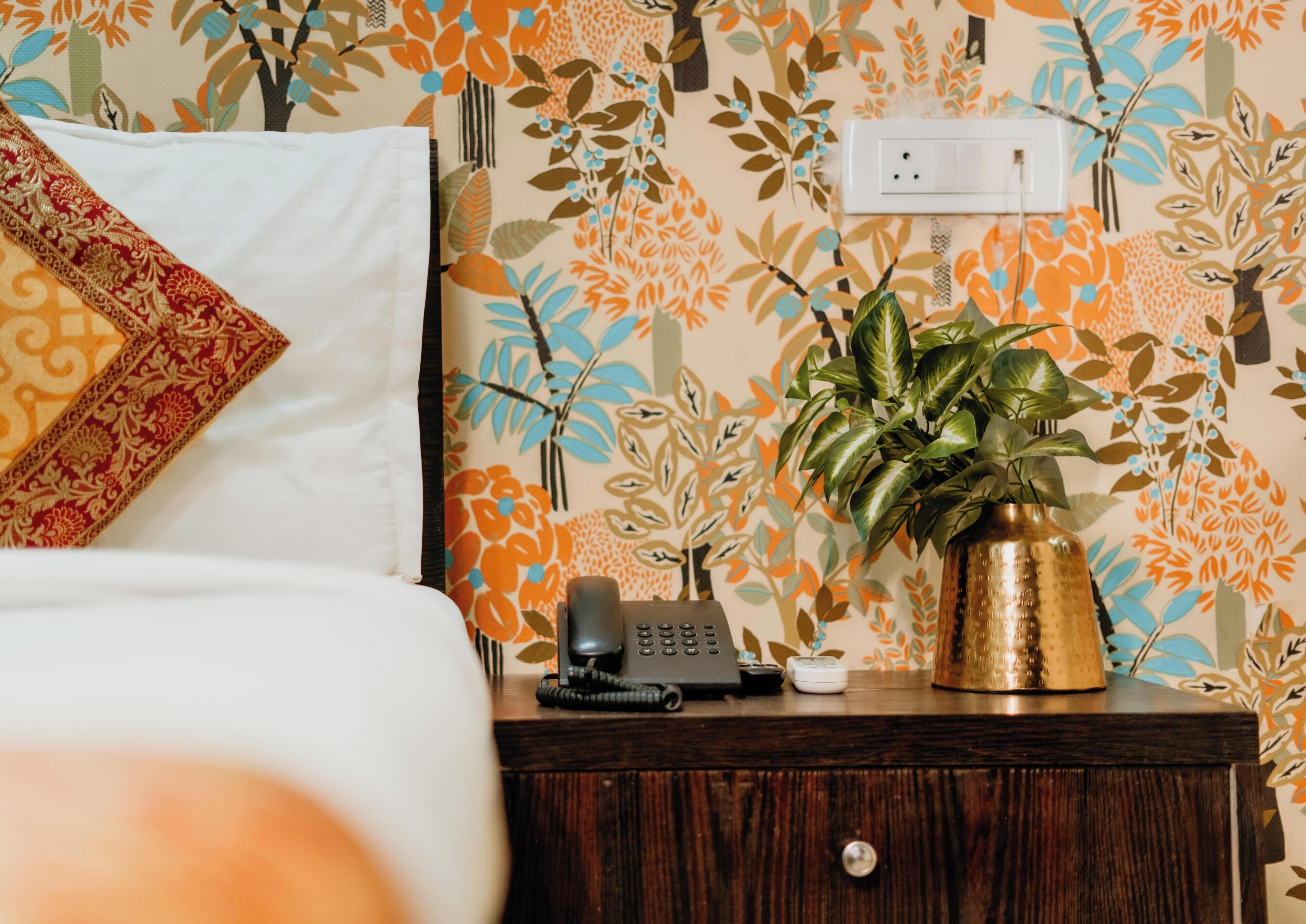 A neatly made bed with a white pillow and a decorative throw, a bedside table with a telephone, a small white object, and a gold vase with a plant.