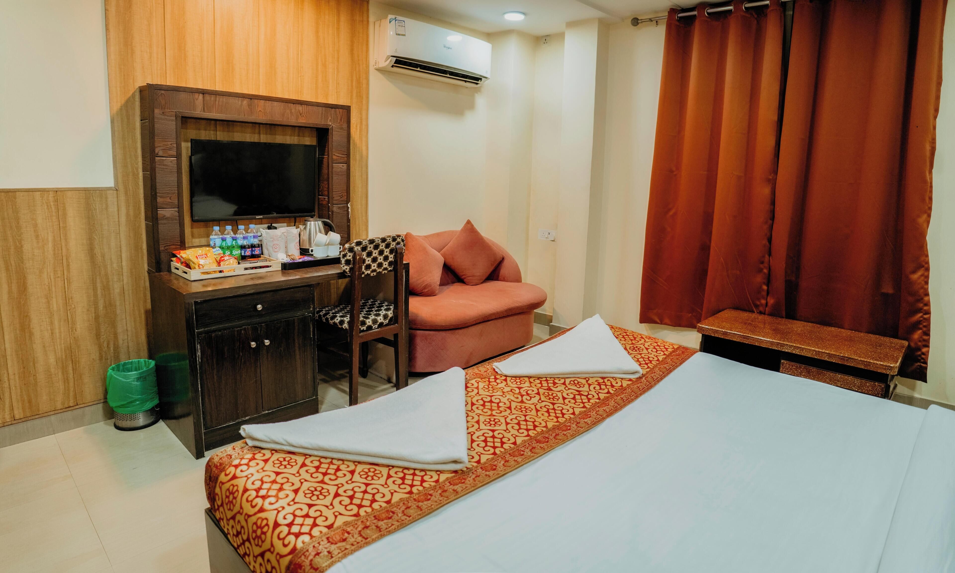 A hotel room with a bed, a desk with a TV, a chair, a sofa, and a window with curtains.