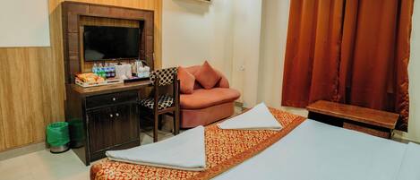 A hotel room with a bed, a desk with a TV, a chair, a sofa, and a window with curtains.