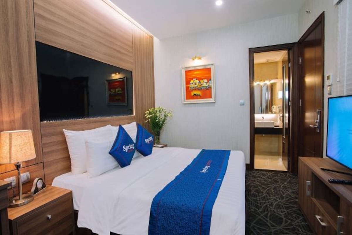 Habitación de hotel con una cama, televisión, escritorio con lámpara y baño visible a través de una puerta abierta.