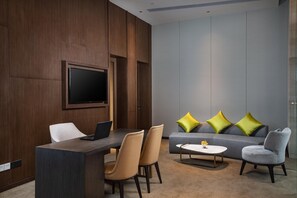 Ein modernes Hotelzimmer mit einer Wand aus Holzpaneelen, einem Flachbildfernseher, einem Schreibtisch mit einem Laptop, einem Sofa mit grünen Kissen und einem Couchtisch.