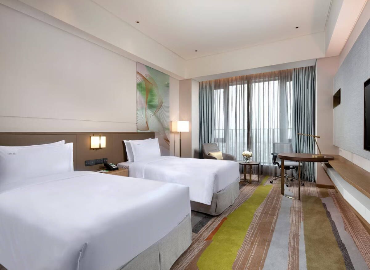A hotel room with two beds, a desk, a chair, a lamp, and a large window with curtains.