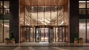 Entrada de um edifício moderno com porta giratória, grandes painéis de vidro e plantas em vasos.